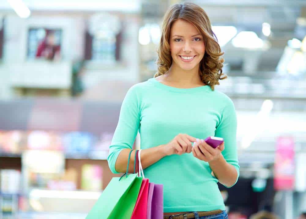 Woman holding shopping bags and using a smartphone, standing indoors in a brightly lit store or mall.